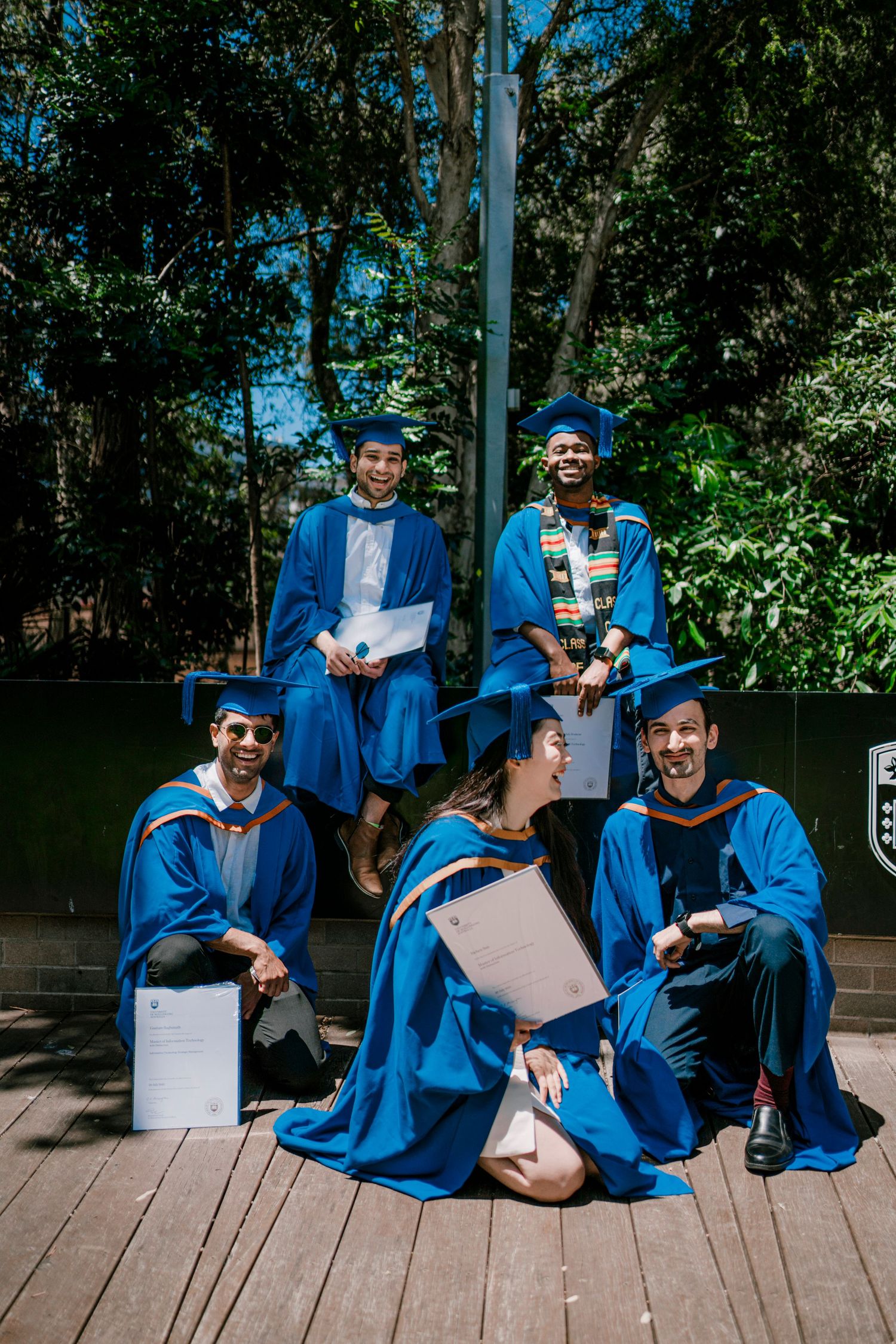 Graduates in blue academic regalia pose together on wooden platform during celebration.