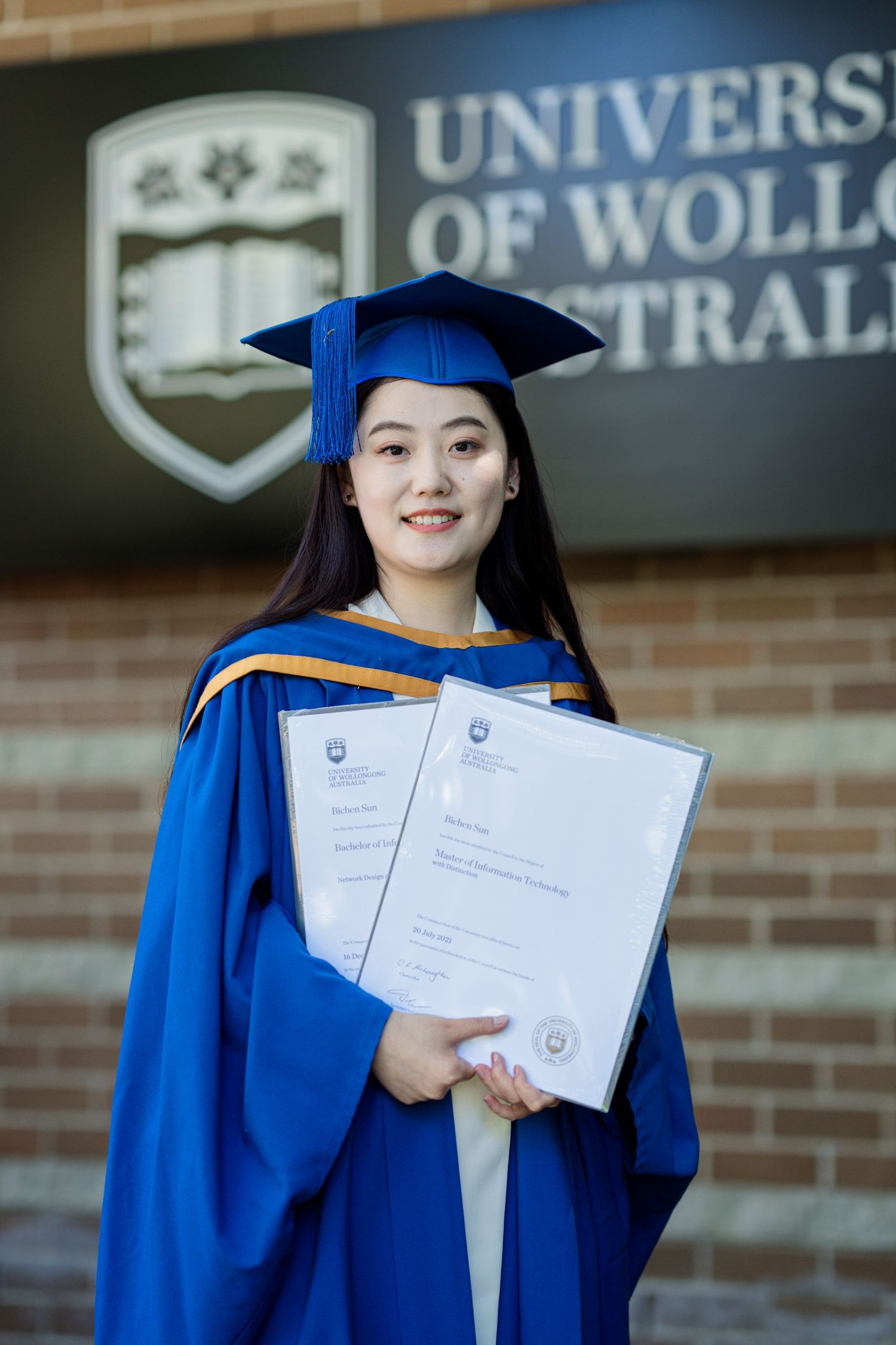Graduate in blue academic regalia holding diploma in front of University of Wollongong sign.
