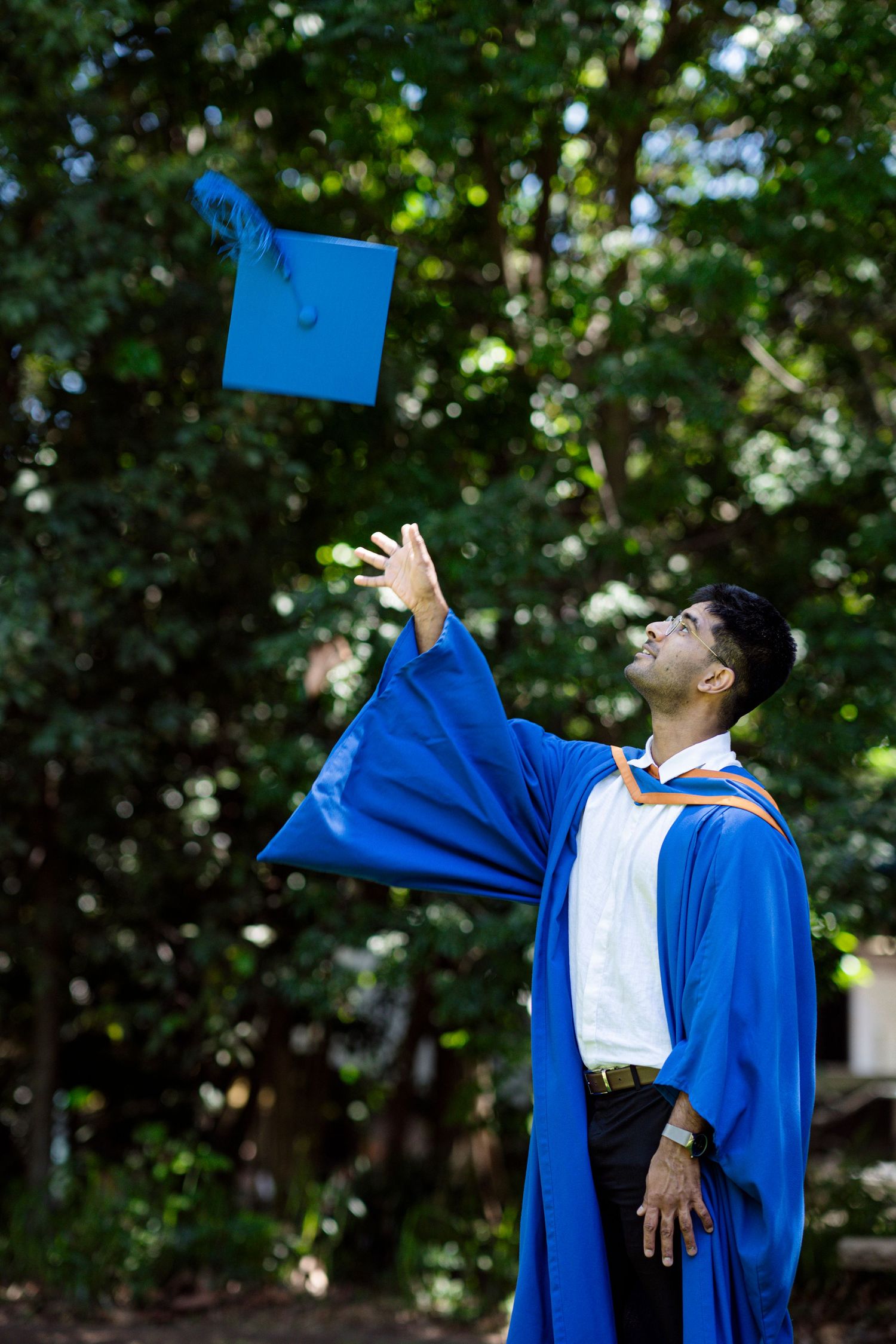 Graduate in blue regalia tosses graduation cap into the air against a leafy green background.