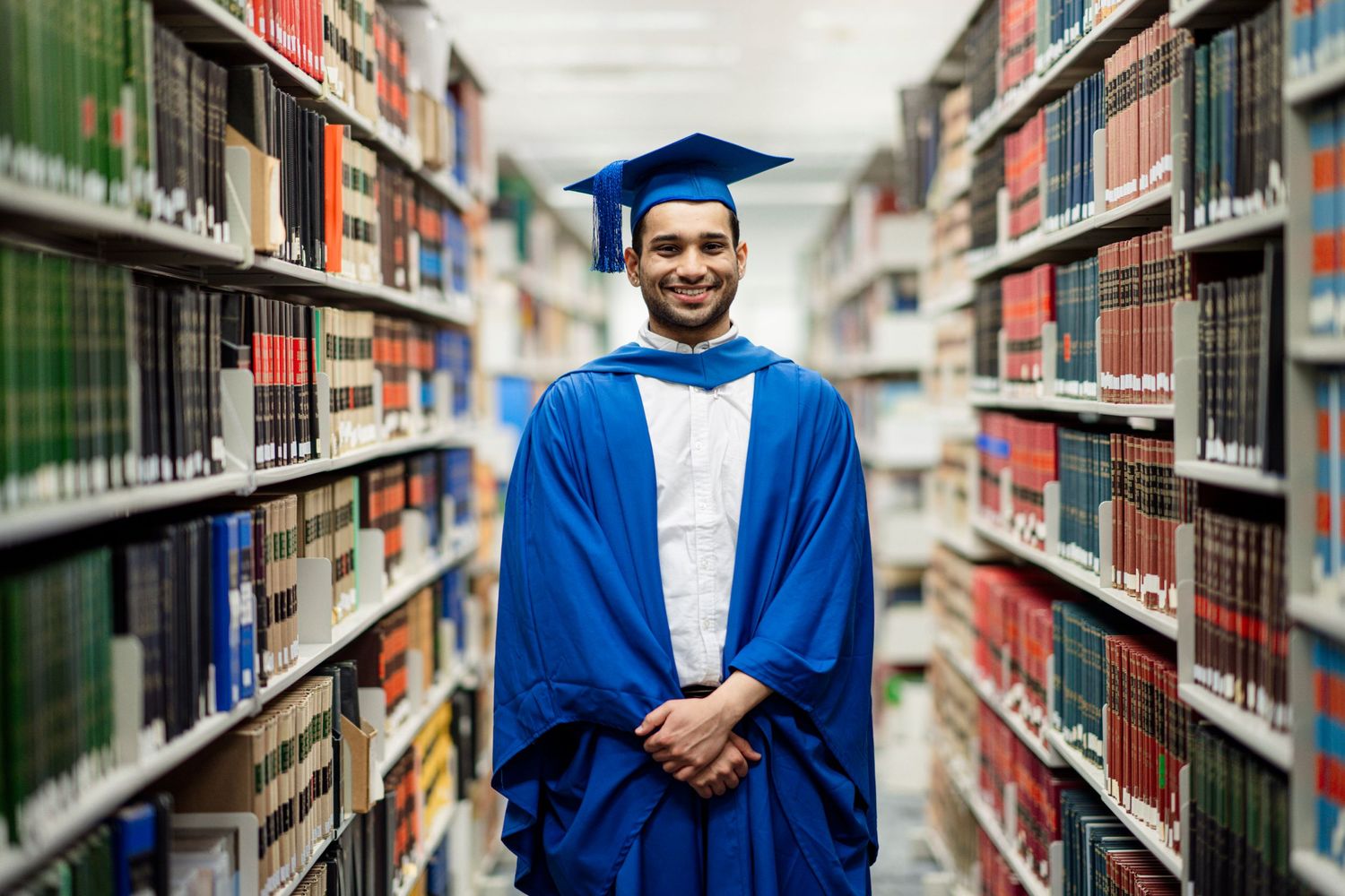 Graduate in academic regalia smiles while standing in university library aisle.