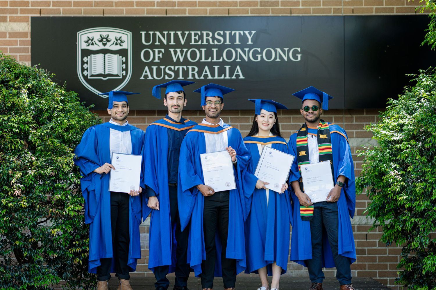 Graduates in blue robes holding diplomas in front of University of Wollongong sign.