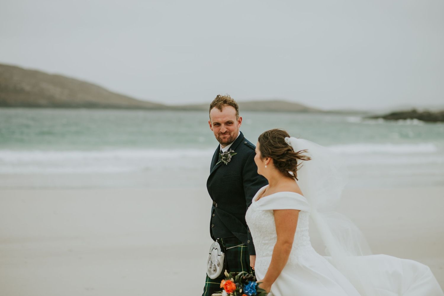 A bride and groom share a moment on a white sandy beach with mountains in the background.