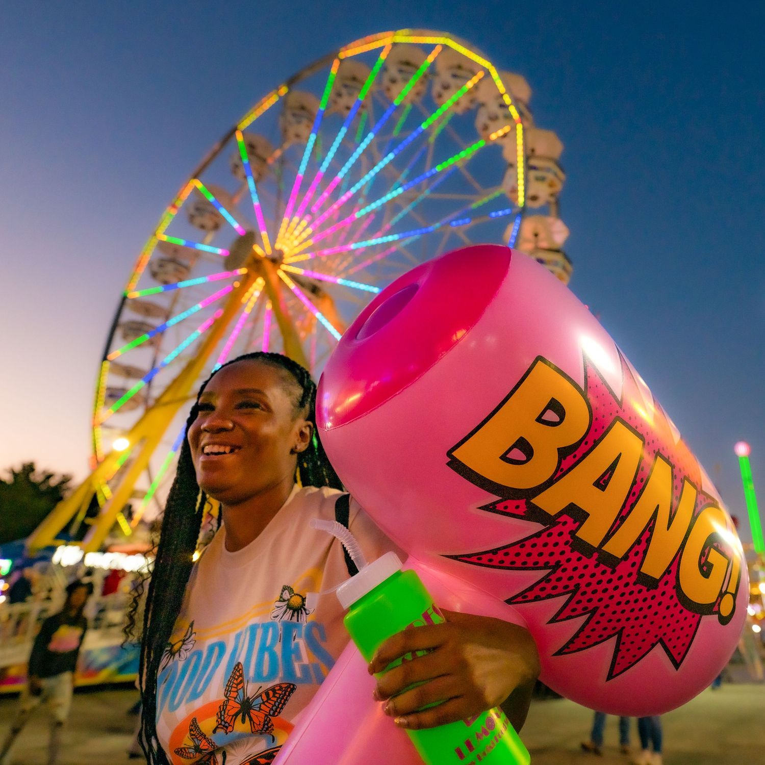 Black women w dreads at the fair with a ferris wheel behind. Smiling big & holding a large pink inflatable toy mallet