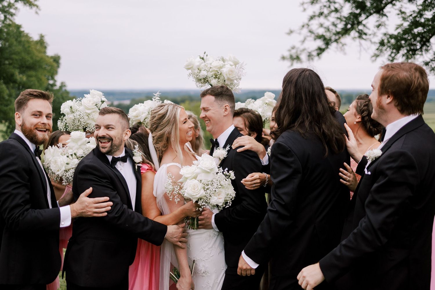 Wedding party celebrates with joyful hugs and laughter outdoors.