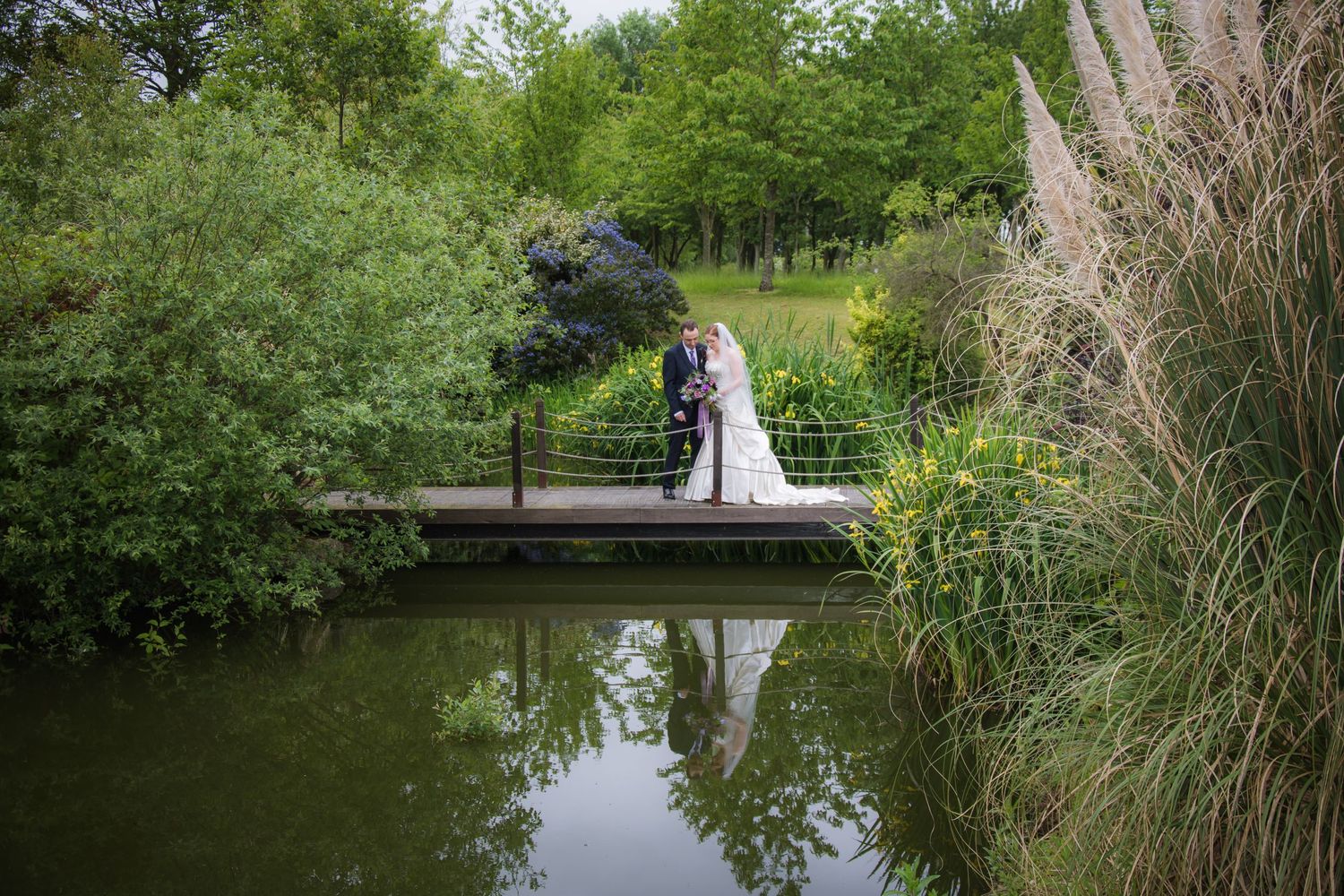 Newlyweds walking across wooden bridge surrounded by lush greenery and pond.