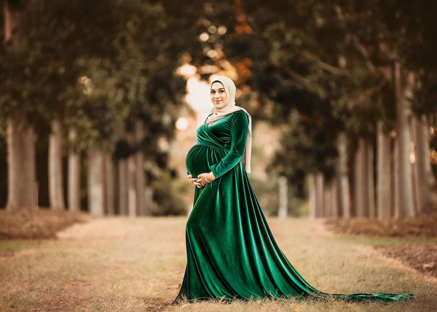 A maternity portrait in an elegant emerald green dress along a tree-lined autumn path.