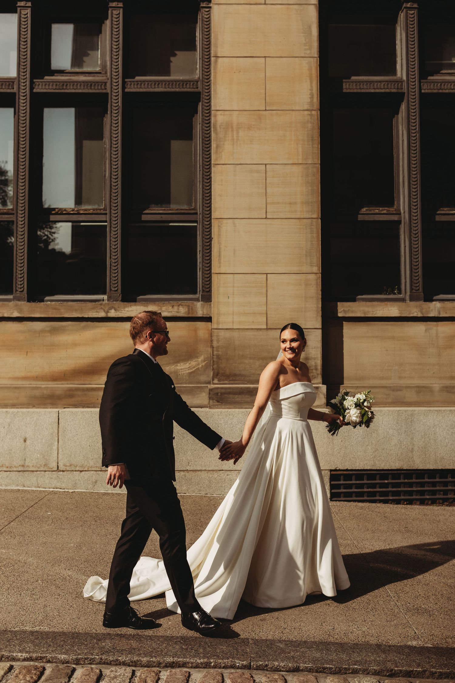 Elegant bride and groom walking outside stone building in dramatic sunlight.