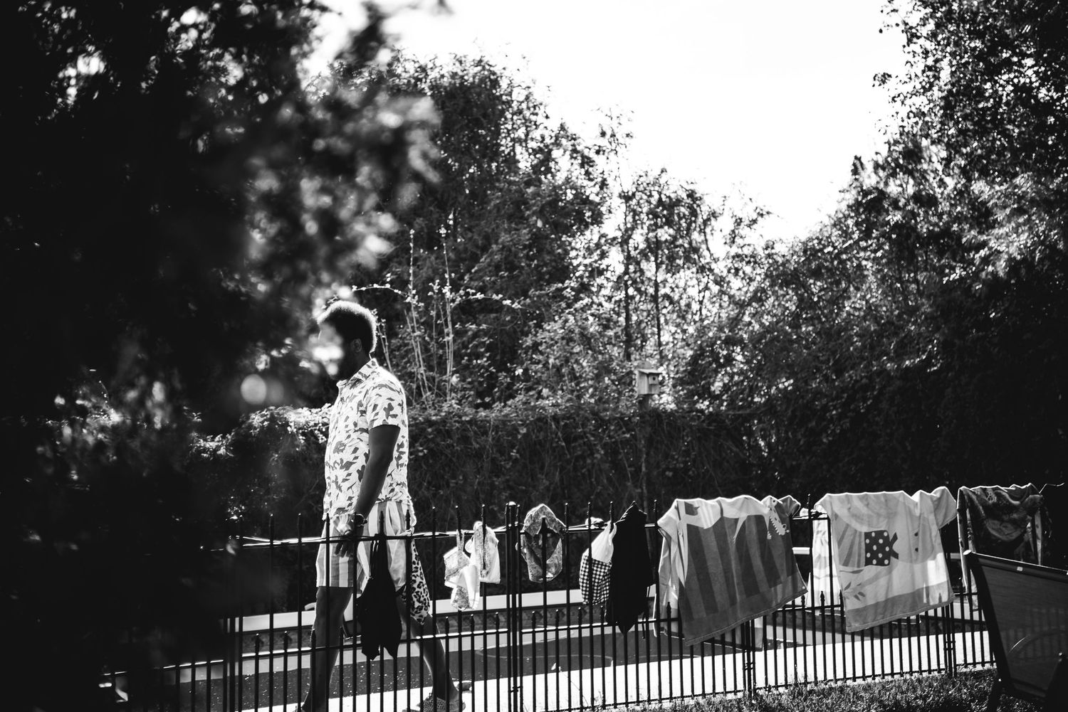 BW image of a black man walking on the other side of a pool fence, his face is covered by a tree branch.