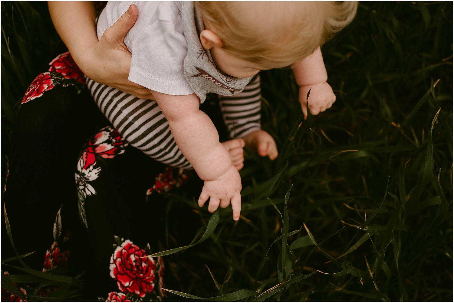 Baby's hands reach toward grass while wearing striped pants and sitting on a floral print.
