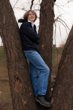 Person in jeans and black jacket climbing and posing playfully between tree trunks.