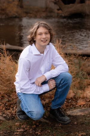 Person in white shirt and jeans crouching on fallen leaves by water.