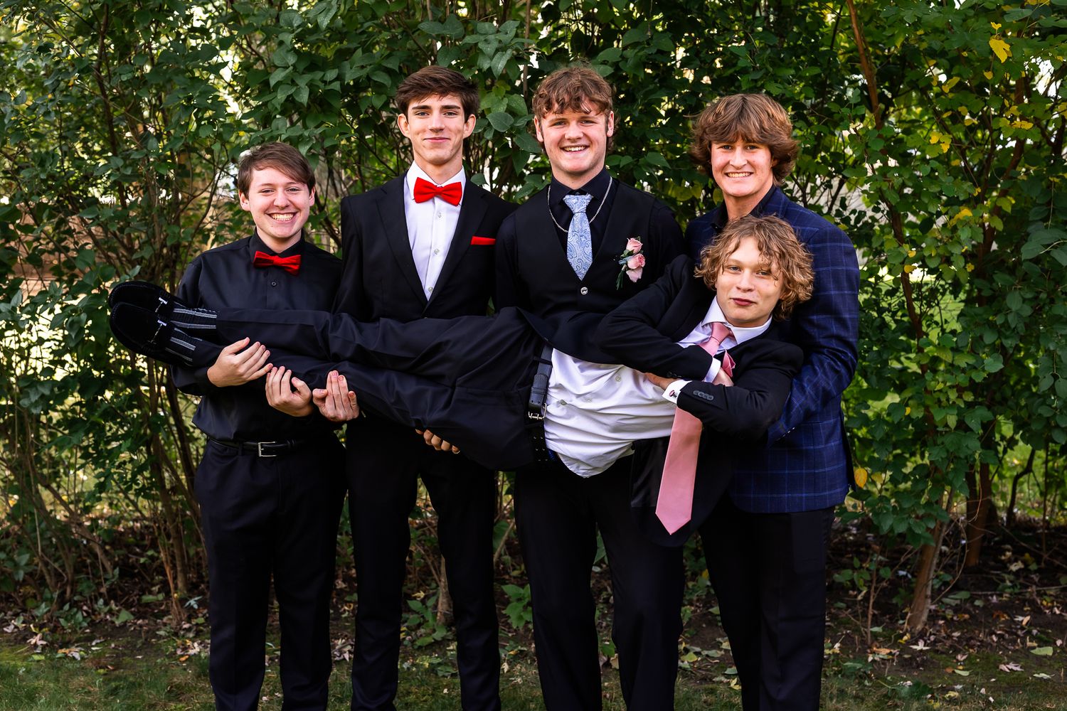 Group of formally dressed people posing together outdoors among green foliage.