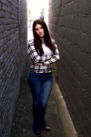 Portrait in urban alley with black brick walls wearing plaid top and jeans.