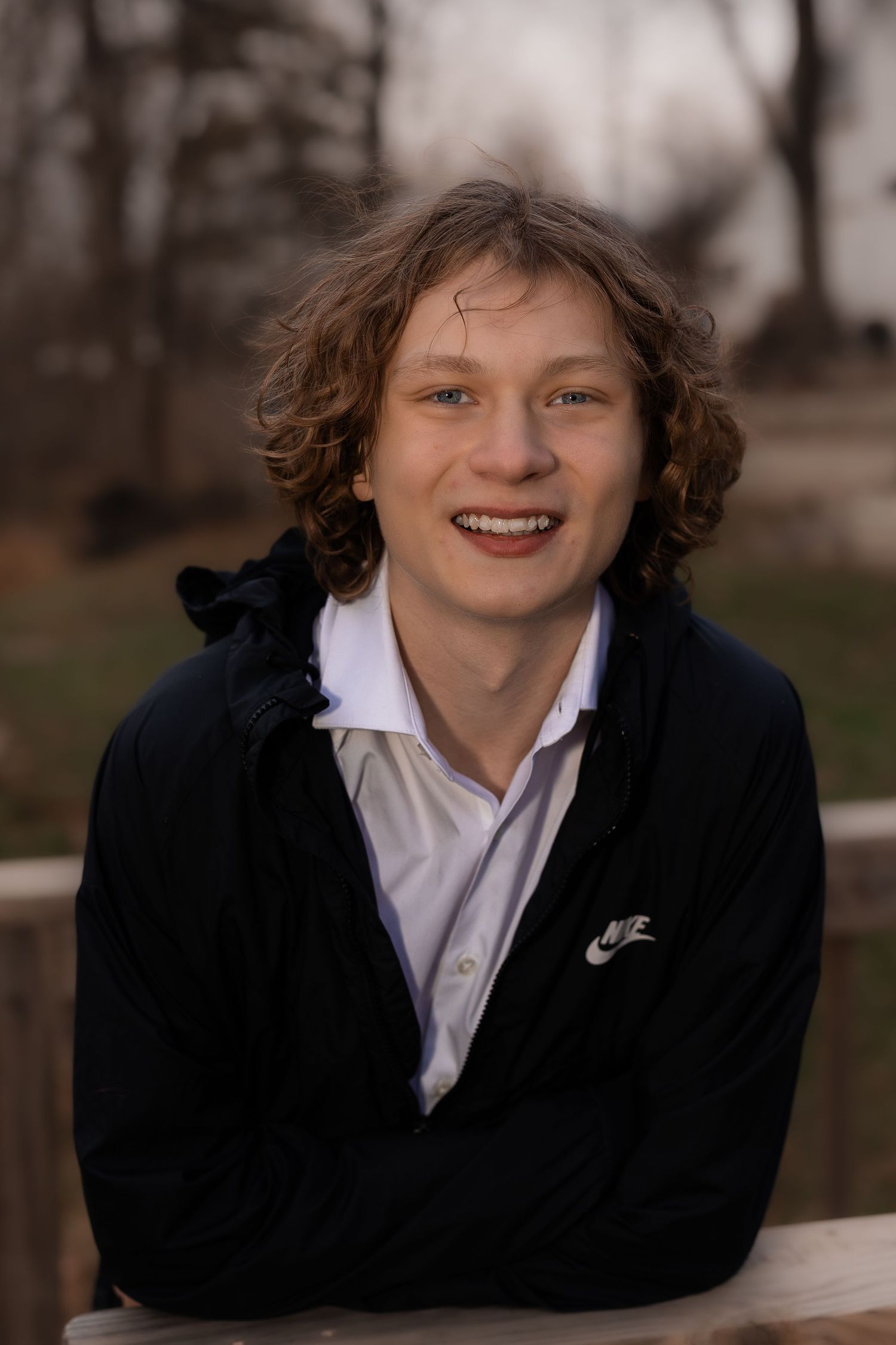 Person wearing black jacket and white shirt smiling warmly against blurred outdoor background.