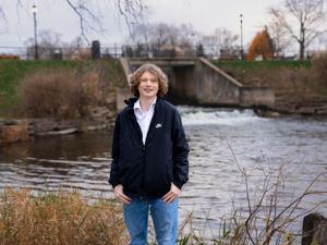 Person in casual wear standing near water with autumn scenery in background.