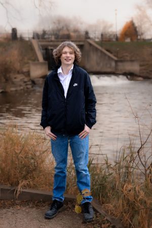 Person in casual attire standing near a waterfront with autumn foliage in the background.