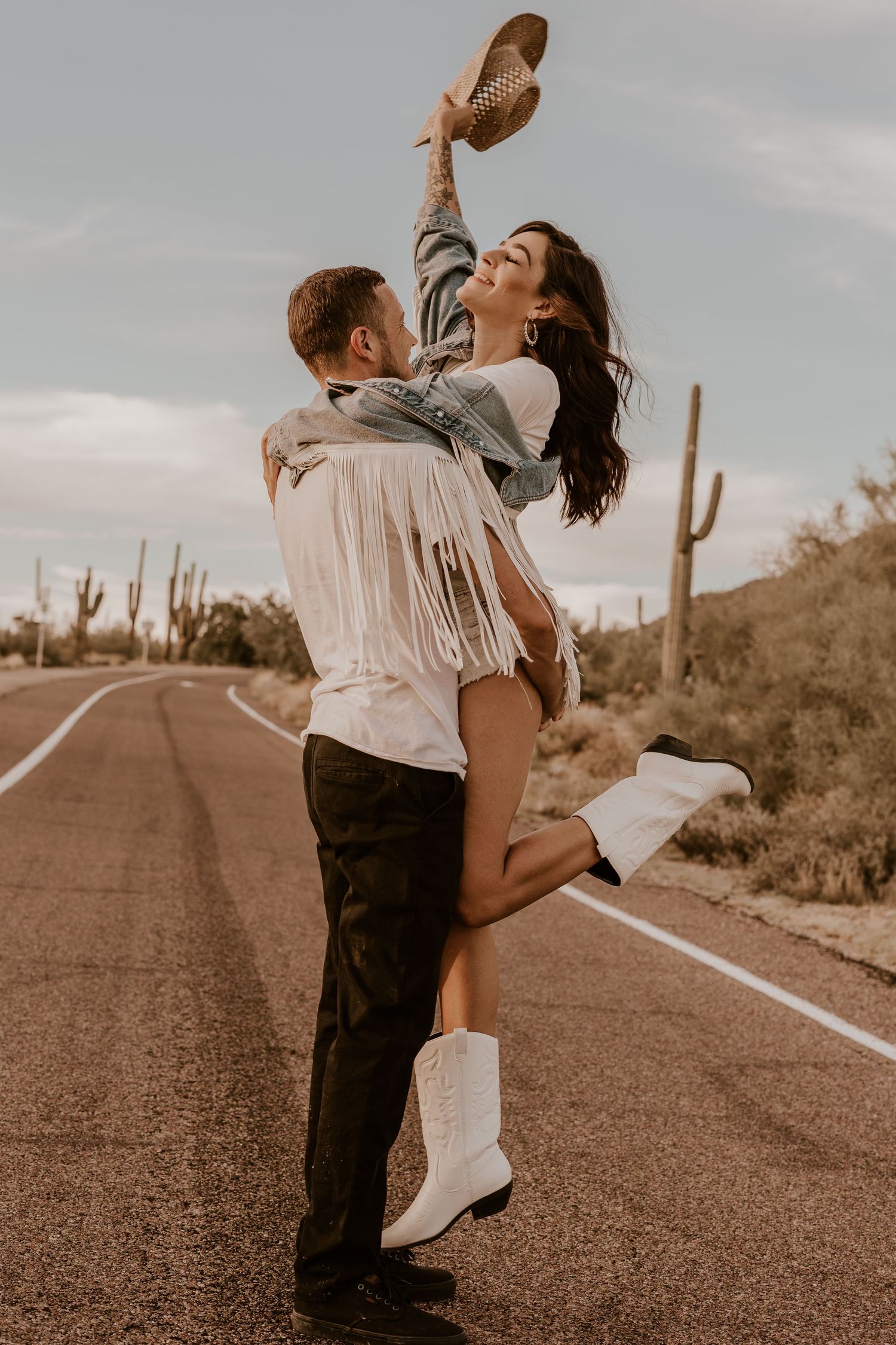 Couple in western attire embracing on desert road with cacti in background.
