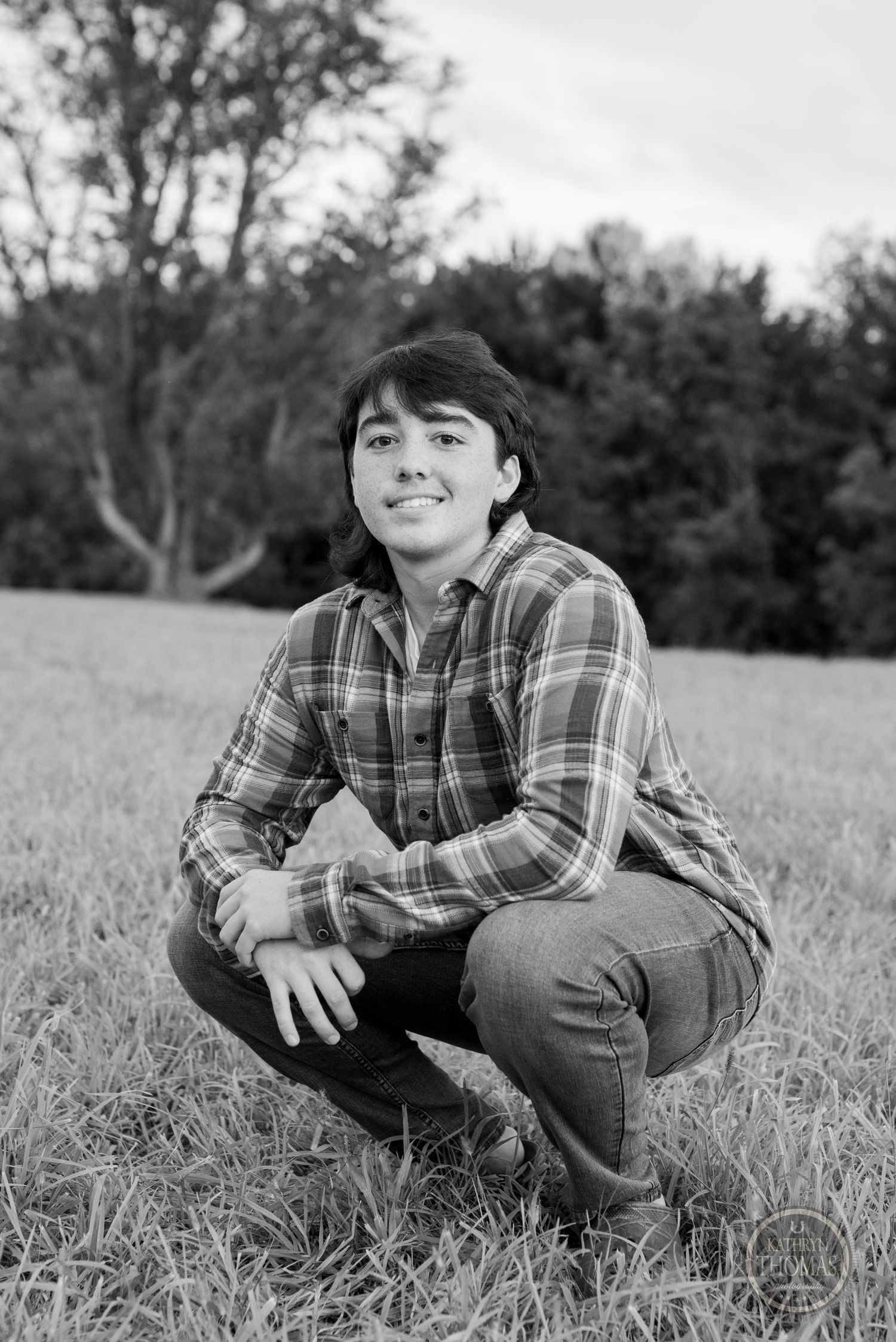 Black and white portrait of person in plaid shirt crouching in grassy field.