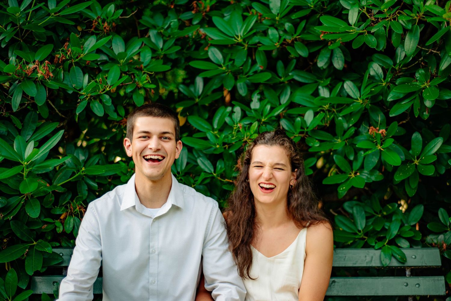 A young couple sits on a bench in the Halifax Public Gardens. They hold hands and are laughing facing the camera.