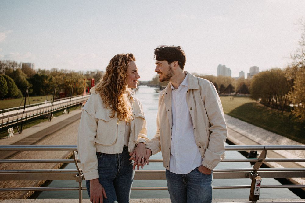 Couple holding hands and gazing at each other on a sunny bridge over a river.