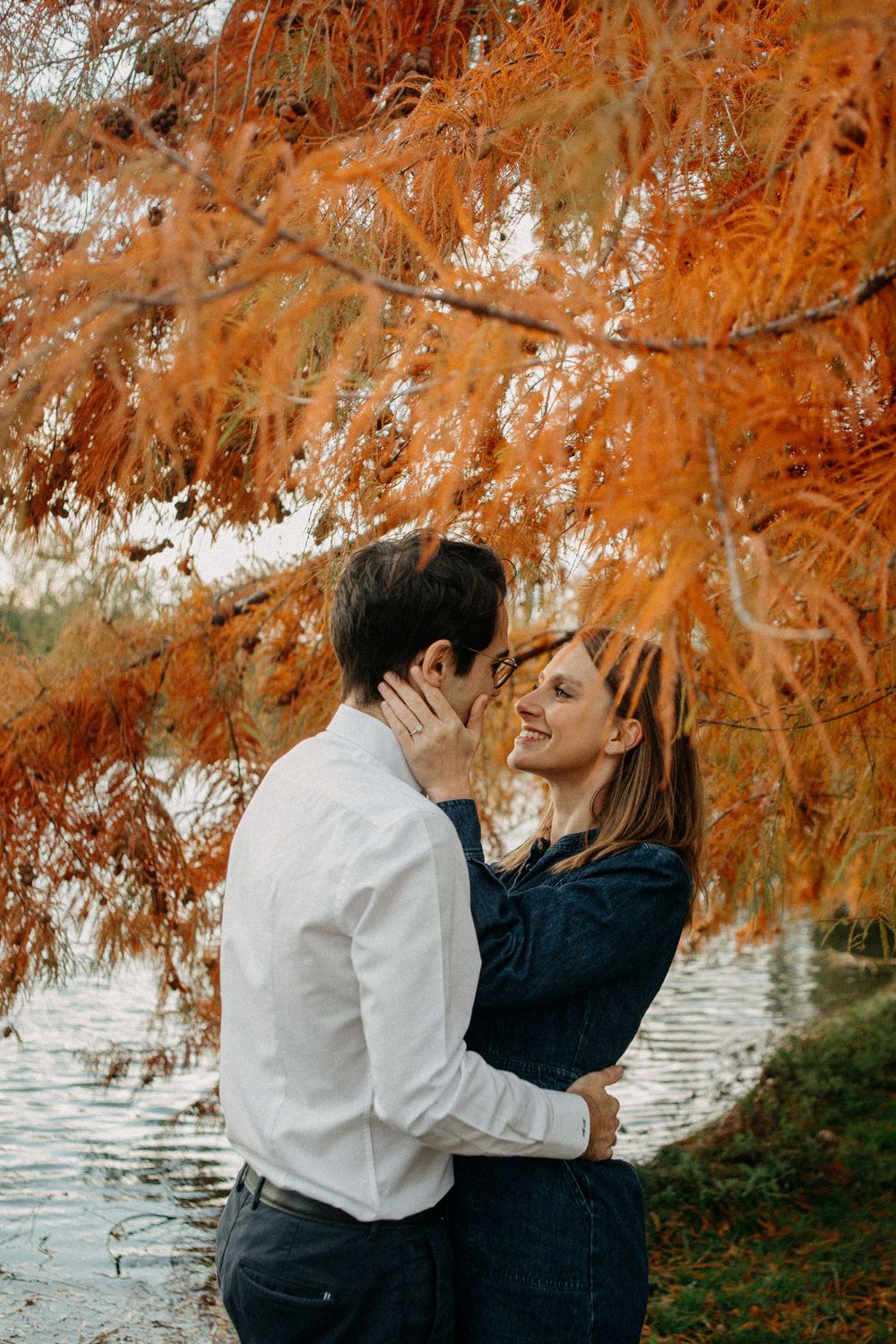Couple sharing a romantic kiss under vibrant orange autumn foliage beside a lake.