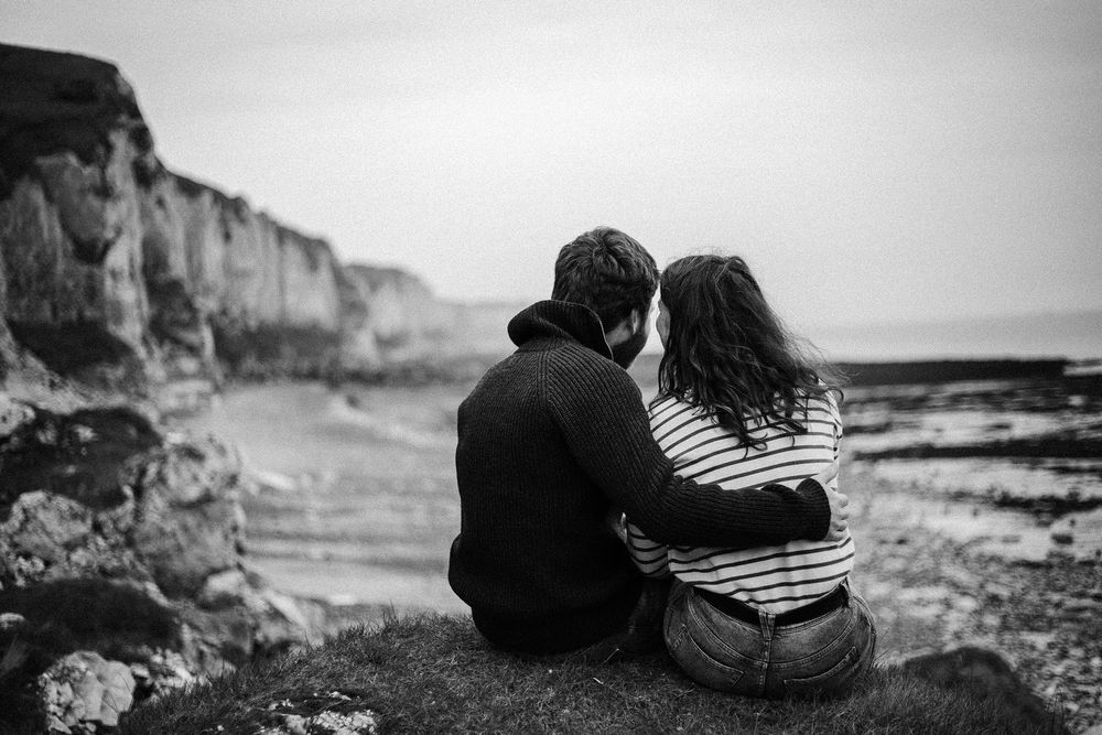 Black and white photo of a couple sitting together overlooking a dramatic coastal cliff beach.