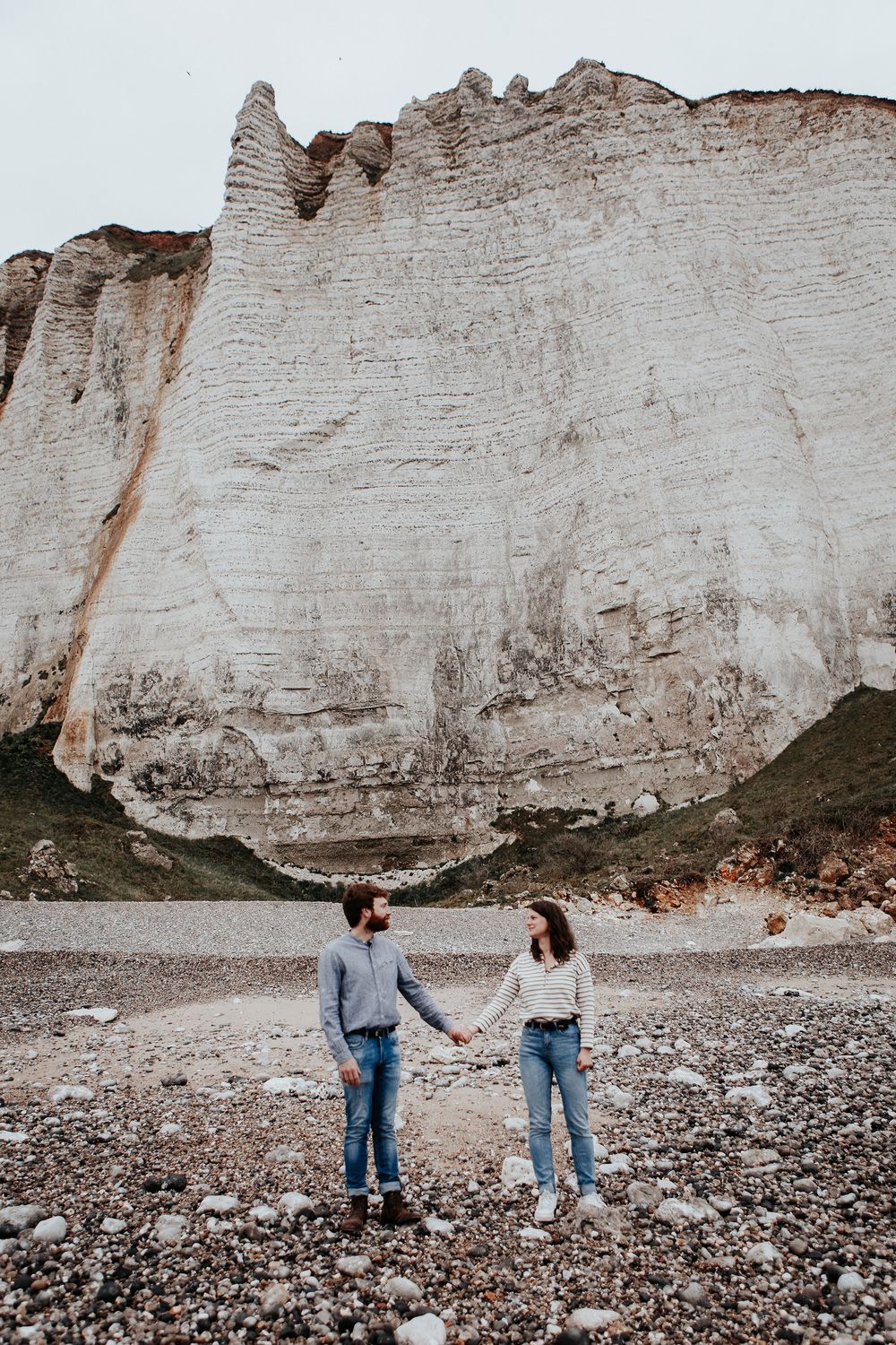 Couple holding hands in front of massive white chalk cliffs on a rocky pebble beach.