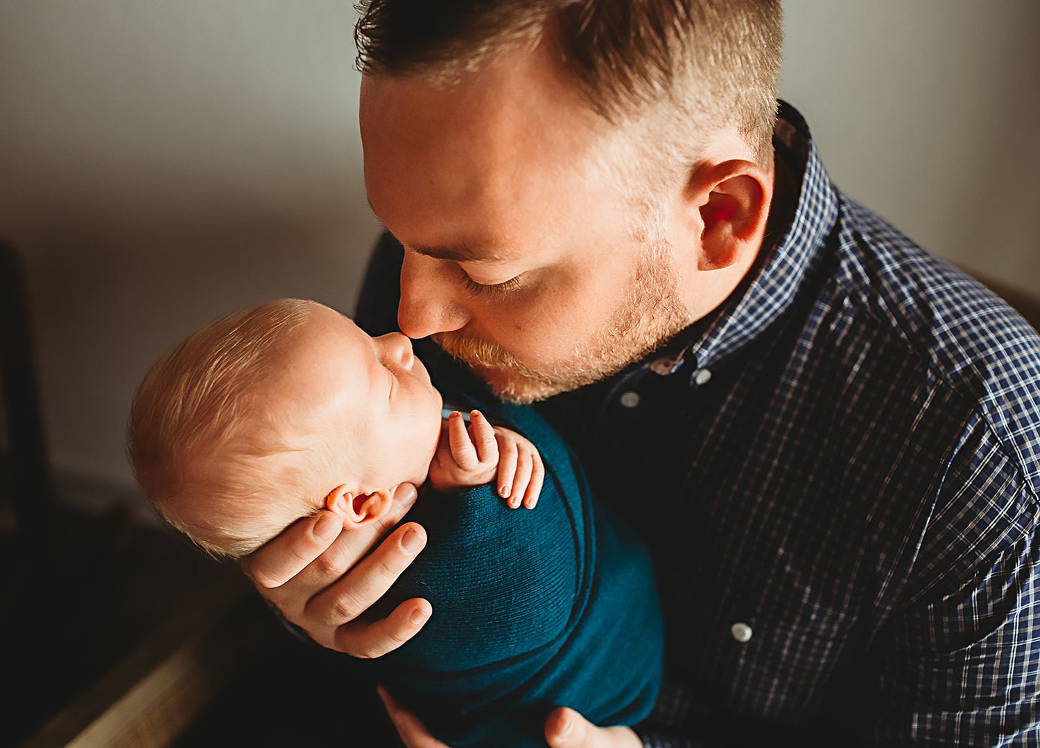 Daddy in a plaid shirt tenderly holding and kissing a baby dressed in teal blue in soft lighting.