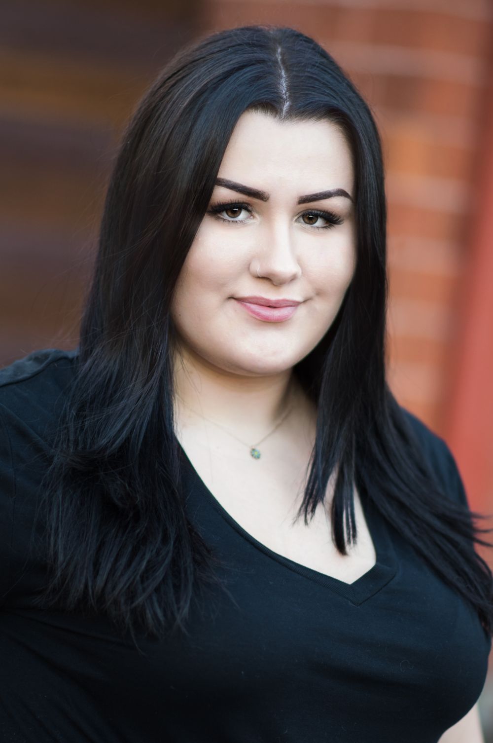 Professional headshot portrait in black v-neck top against brick wall.