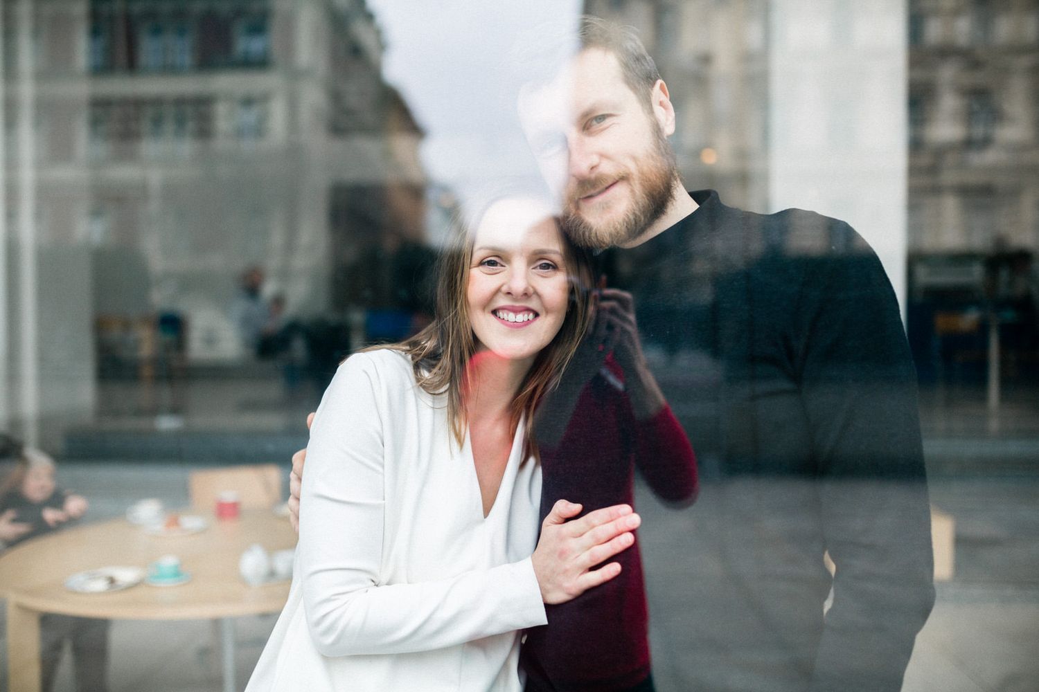 Happy couple embracing and laughing by window reflection in urban setting.