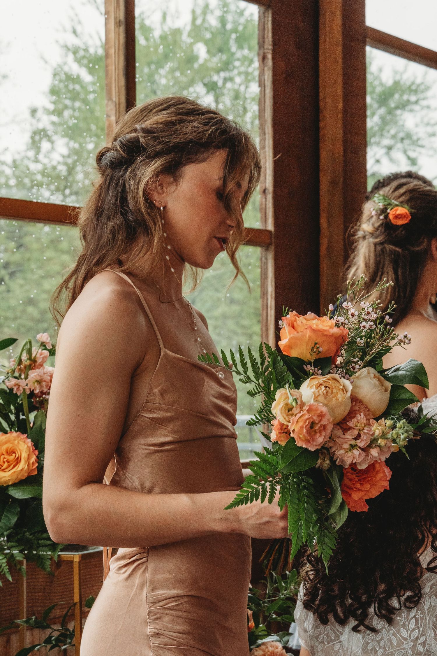 Bridesmaid in copper silk dress holding peach and coral floral bouquet by window.