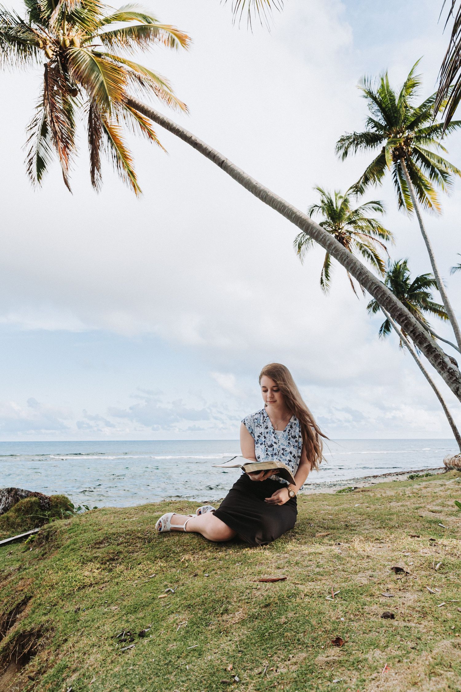Woman reading book under leaning palm trees on tropical coastline.