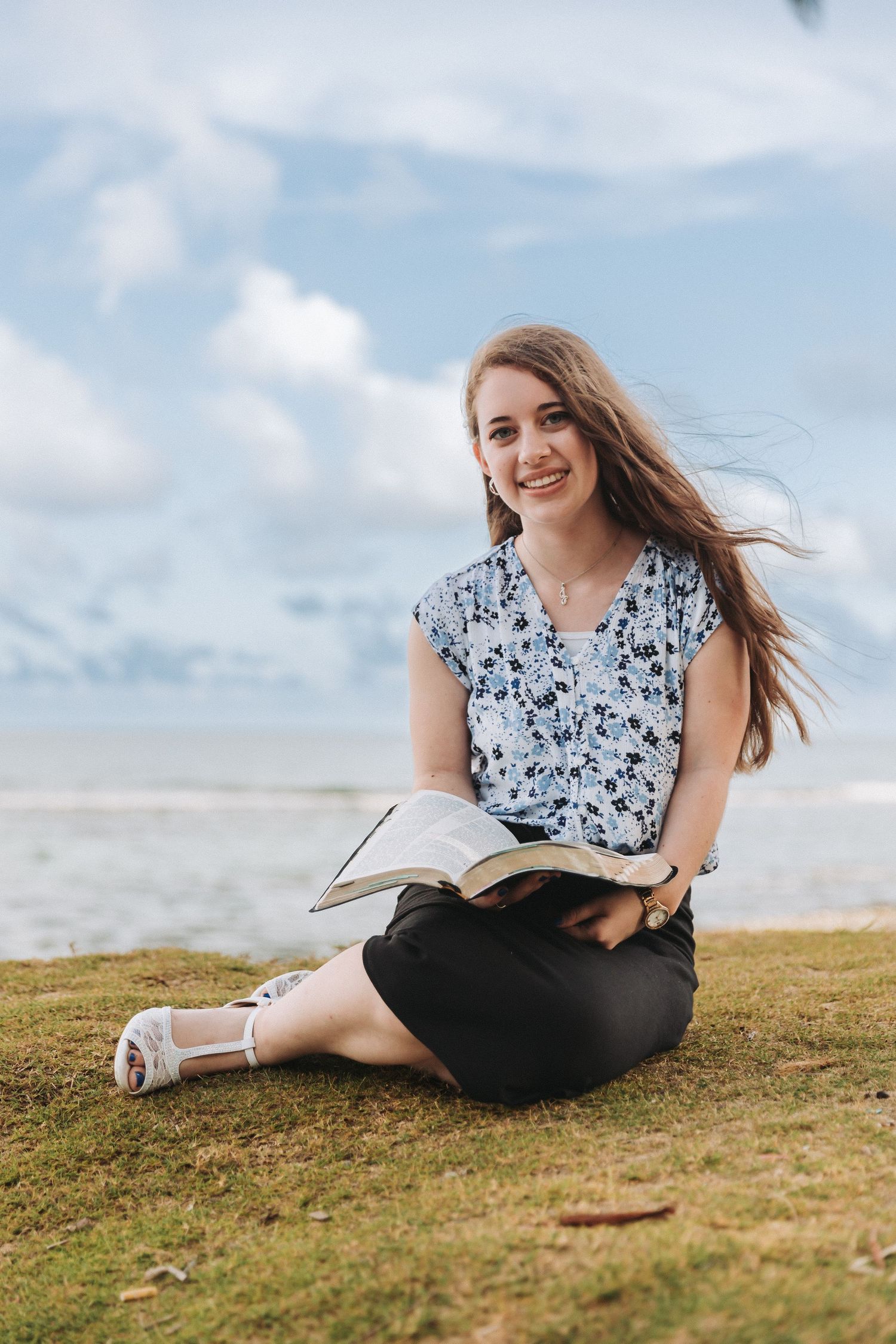 Woman sitting with book on oceanside cliff with palm trees and blue sky.