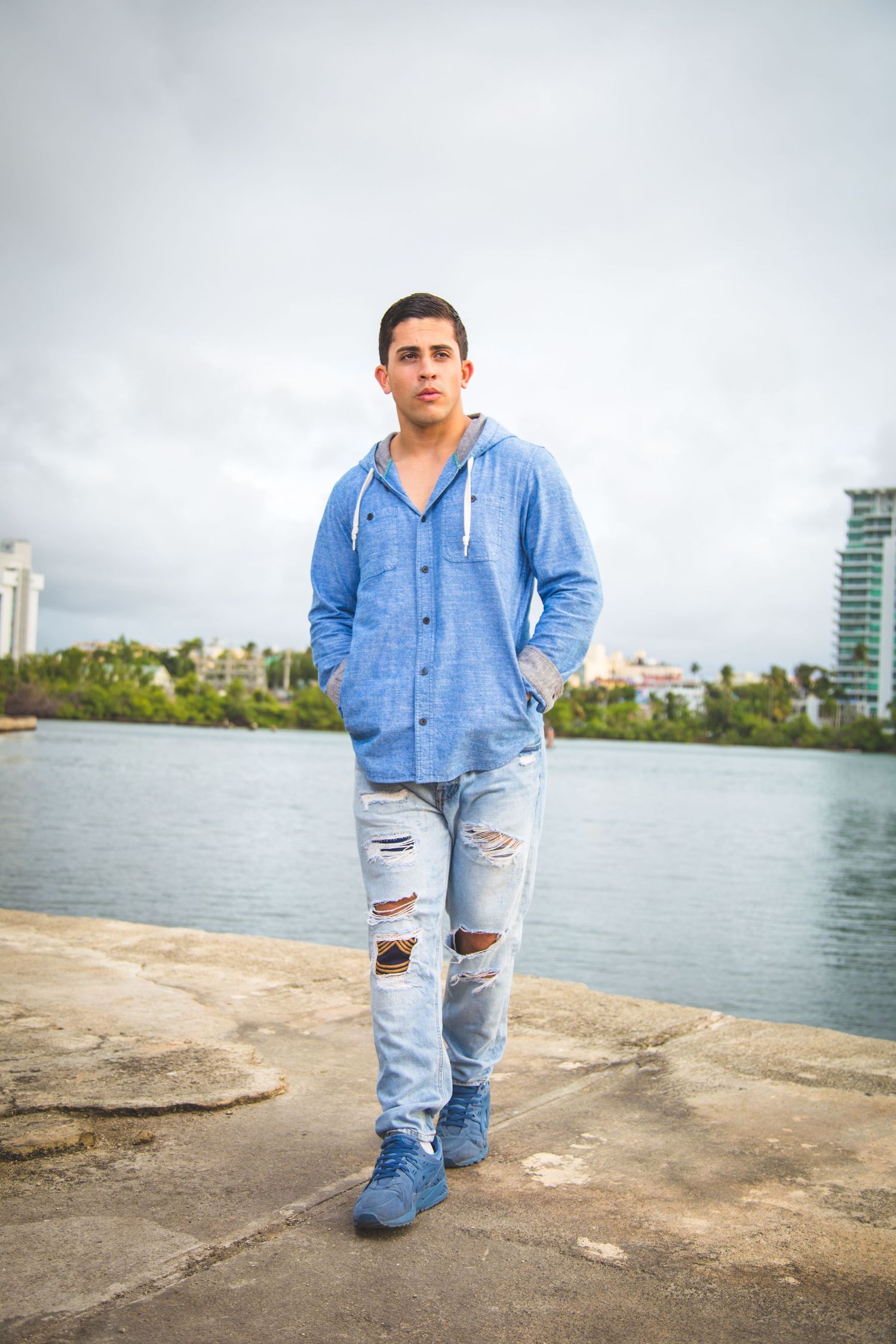 Person in denim jacket and ripped jeans standing by waterfront with city buildings in background.