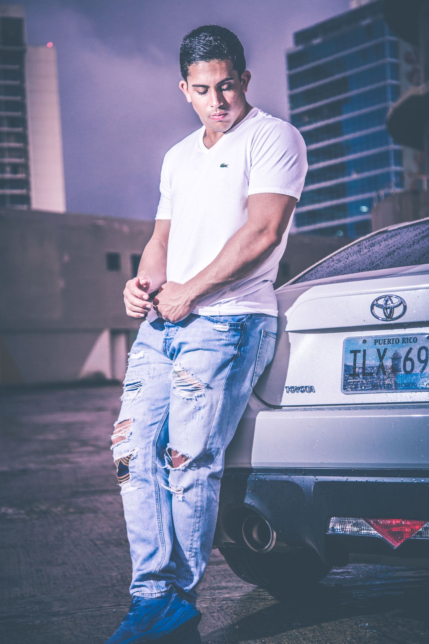 Person in white t-shirt and distressed jeans posing next to white car in urban setting.