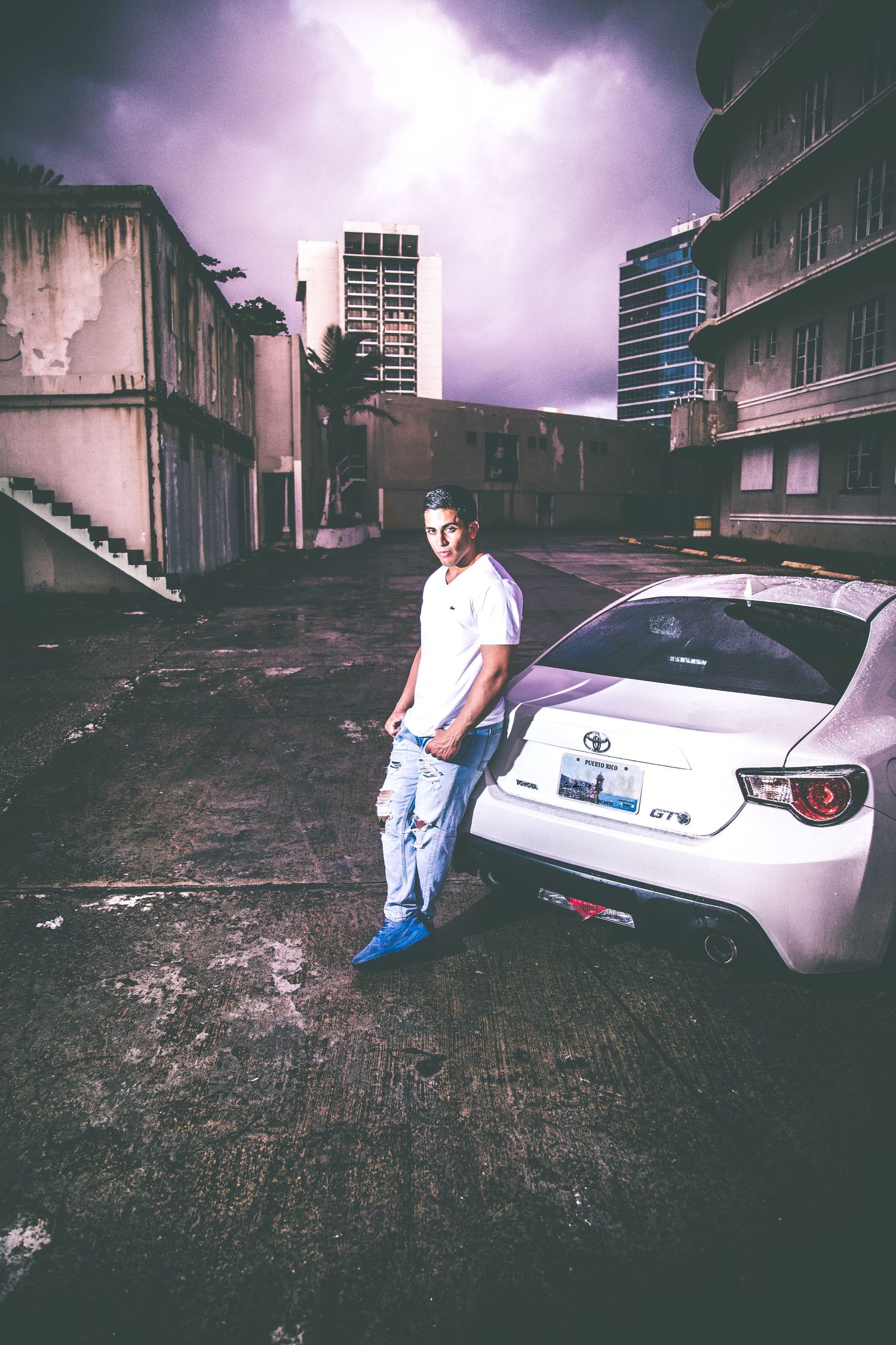 Person leaning against white car with dramatic purple sky and urban buildings in background.