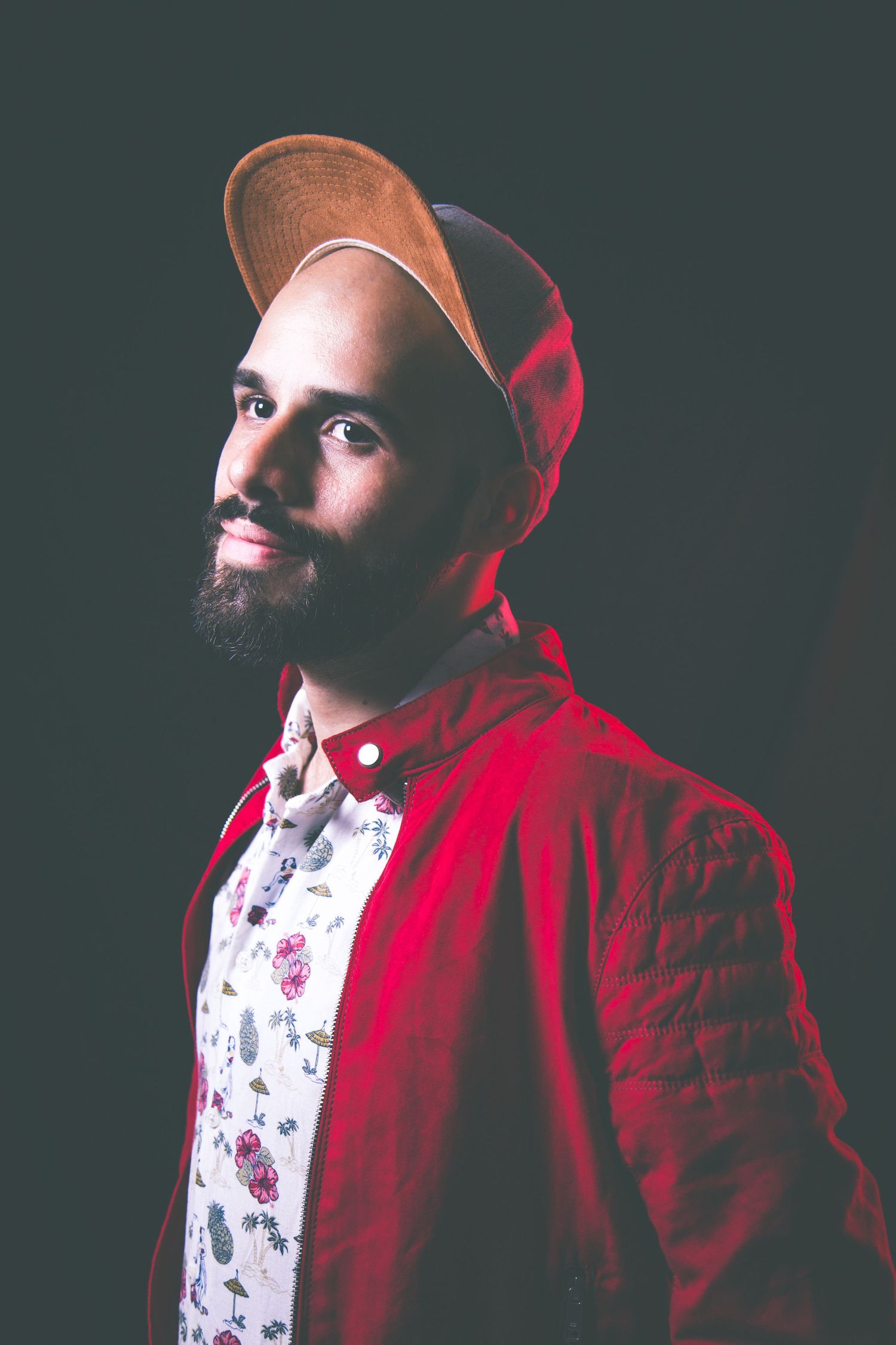 Portrait of someone wearing a red jacket, floral shirt and brown baseball cap against dark background.