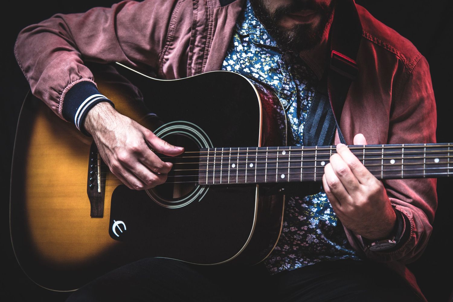Close up detail of hands playing acoustic guitar showing sunburst finish and floral shirt pattern.