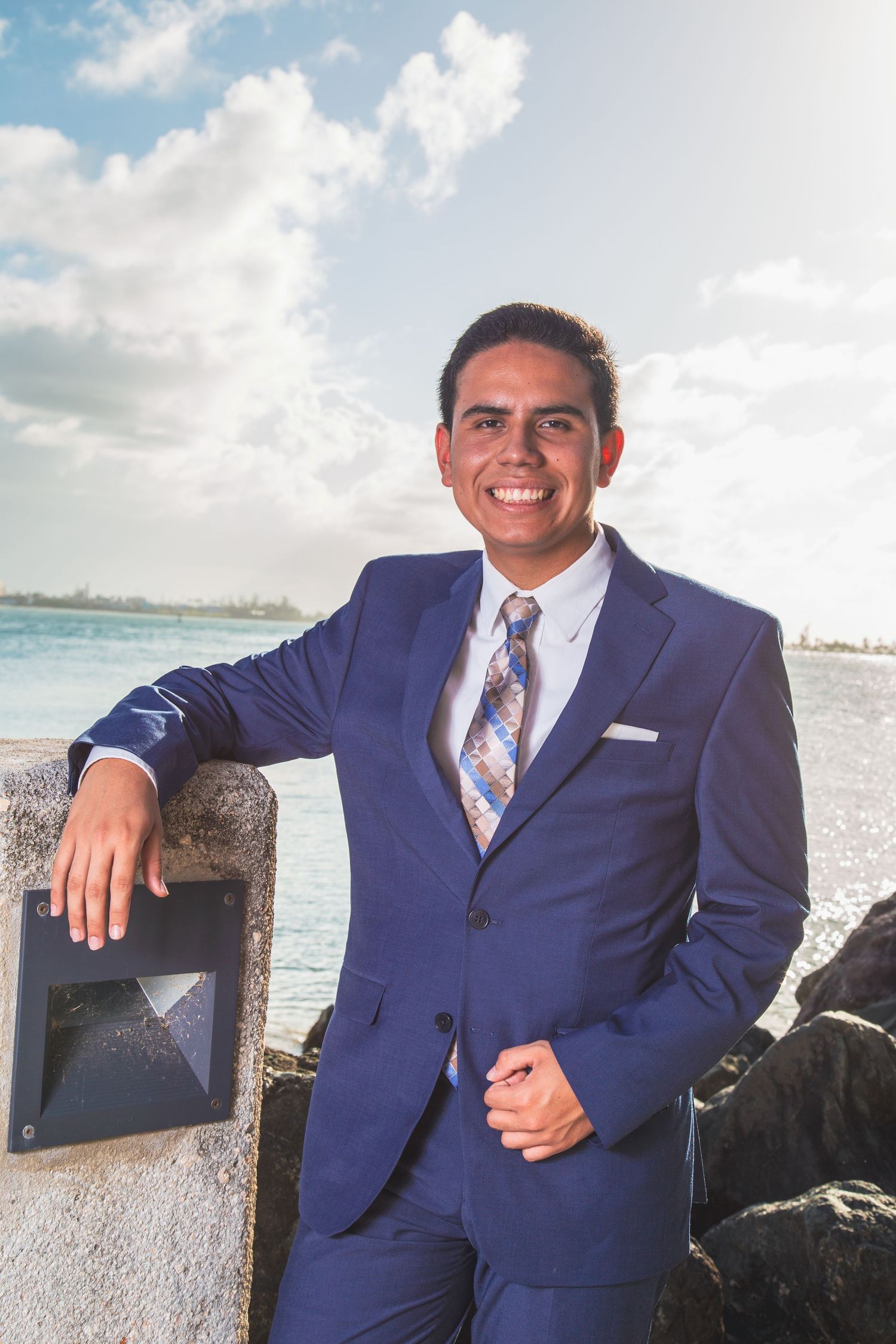 Professional in navy blue suit poses confidently against coastal backdrop on sunny day.