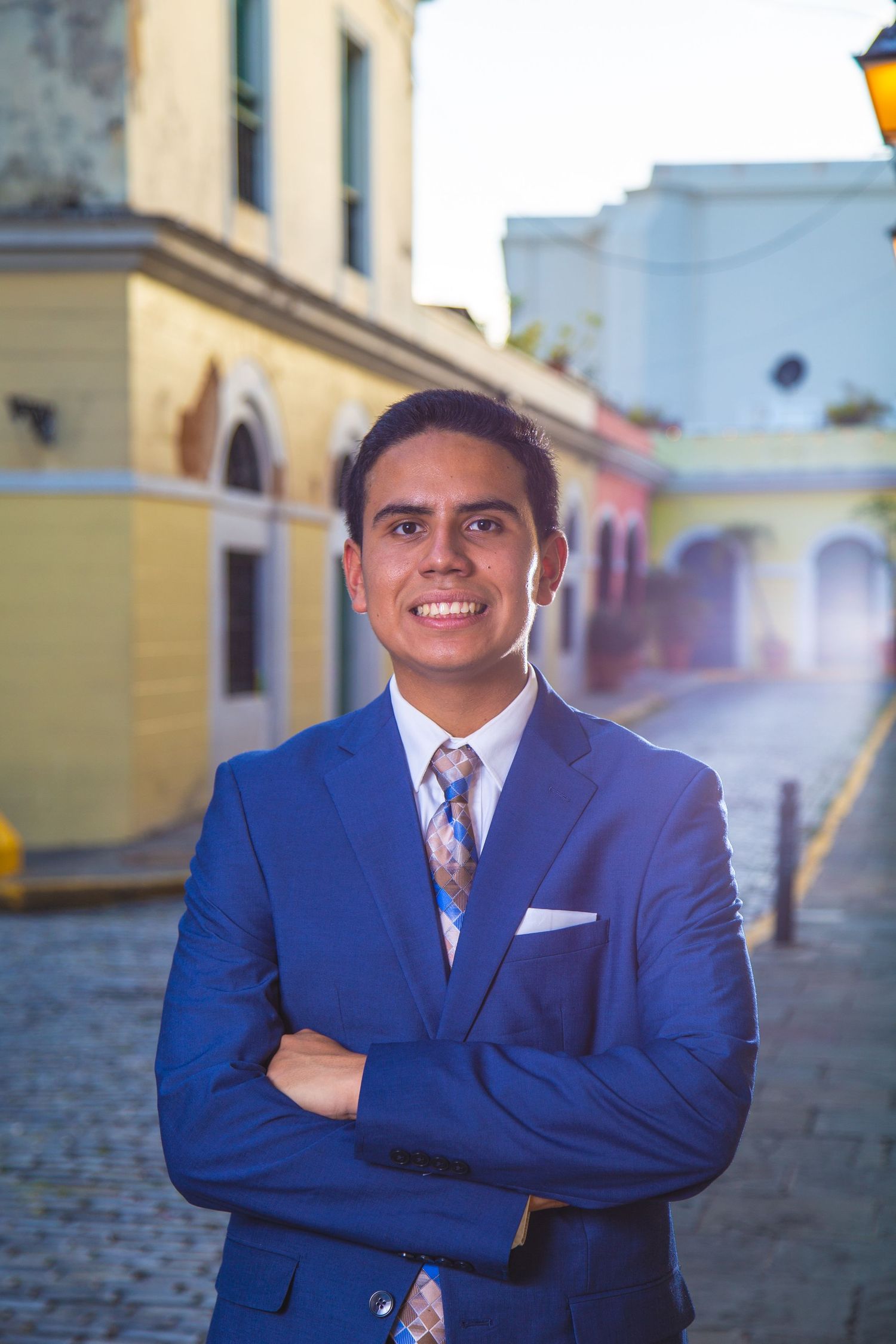 Professional in navy suit stands with crossed arms in colorful colonial street setting.
