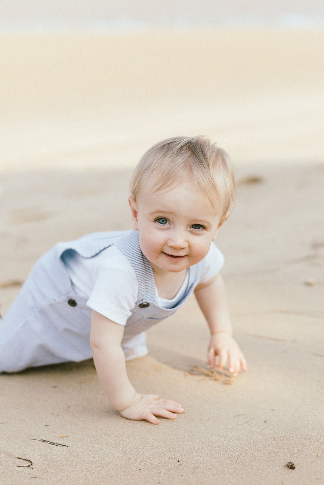 A small child in white clothing lies and smiles while playing in soft sand.