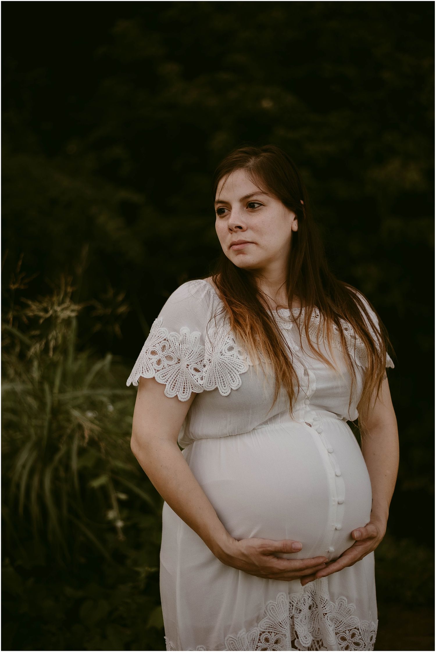 Artistic maternity portrait in a white lace dress against a dark natural background.