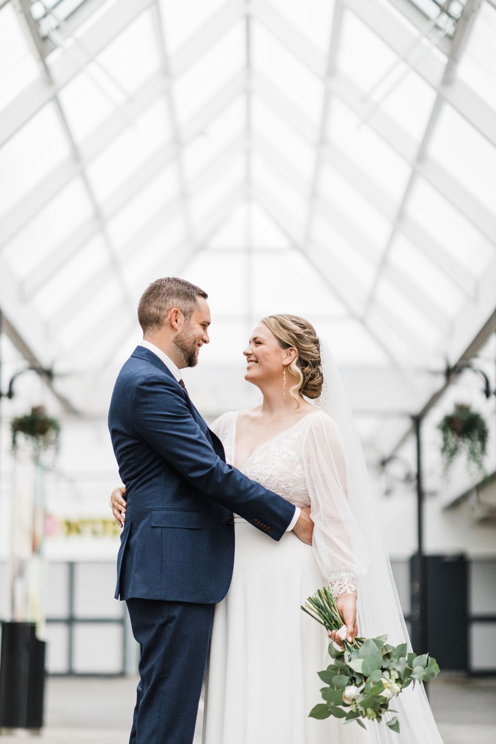 Couple shares joyful moment in elegant wedding attire under a glass conservatory ceiling.