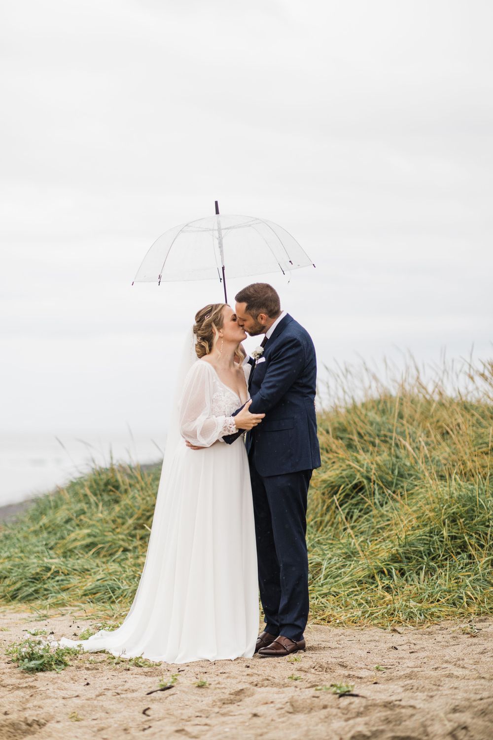 Newlyweds share intimate moment under clear umbrella on coastal cliff.