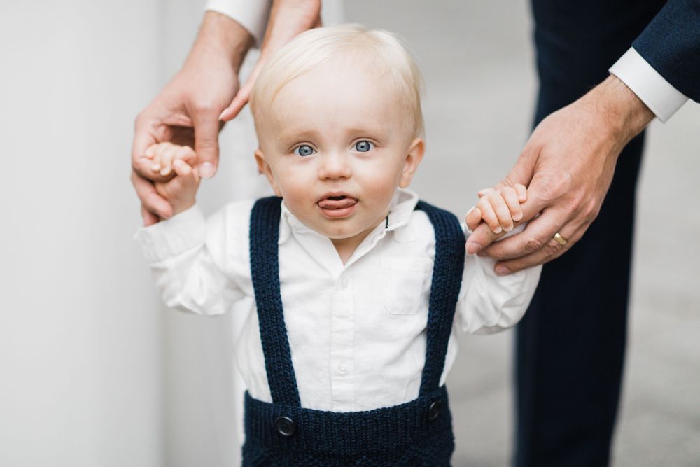 Toddler in white shirt and navy suspenders standing between two people.