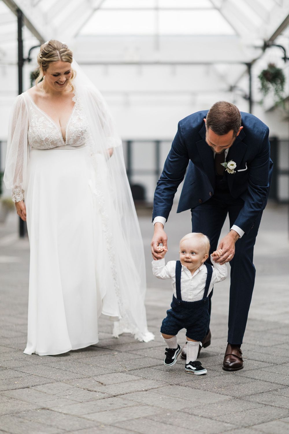 Wedding scene in bright conservatory with couple helping small child take steps.