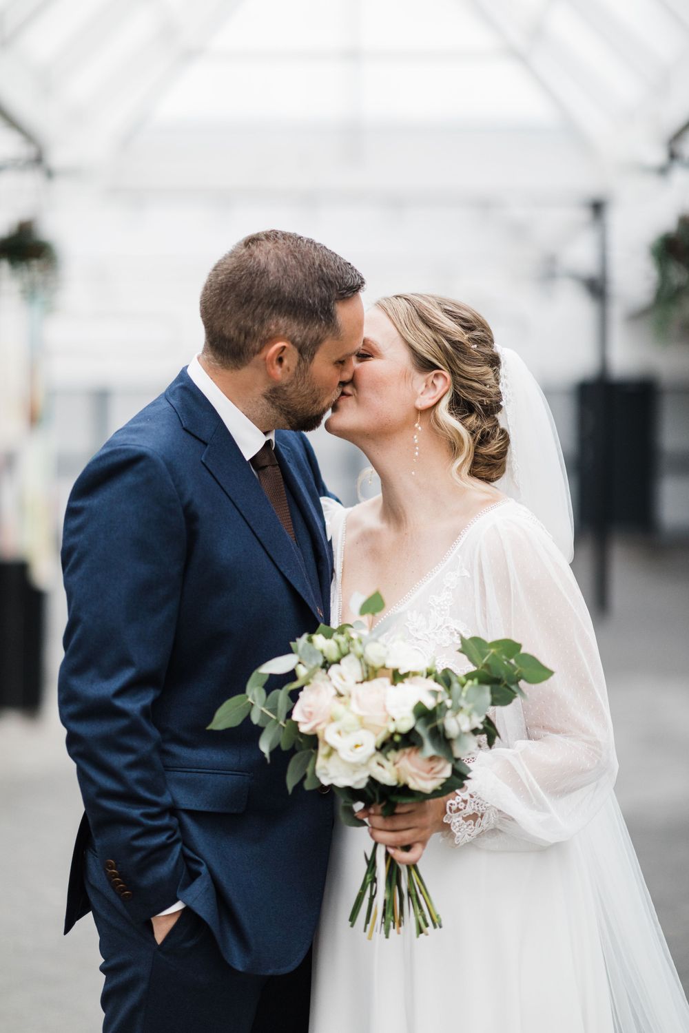 Romantic wedding kiss in an airy conservatory with bride holding white floral bouquet.