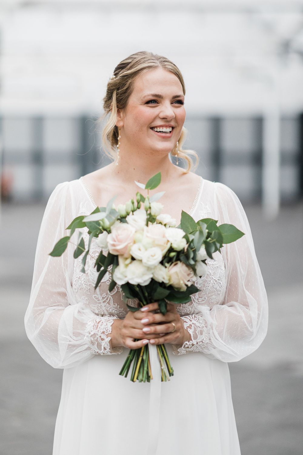 Bride in white lace dress smiles while holding elegant bouquet of white roses and greenery.