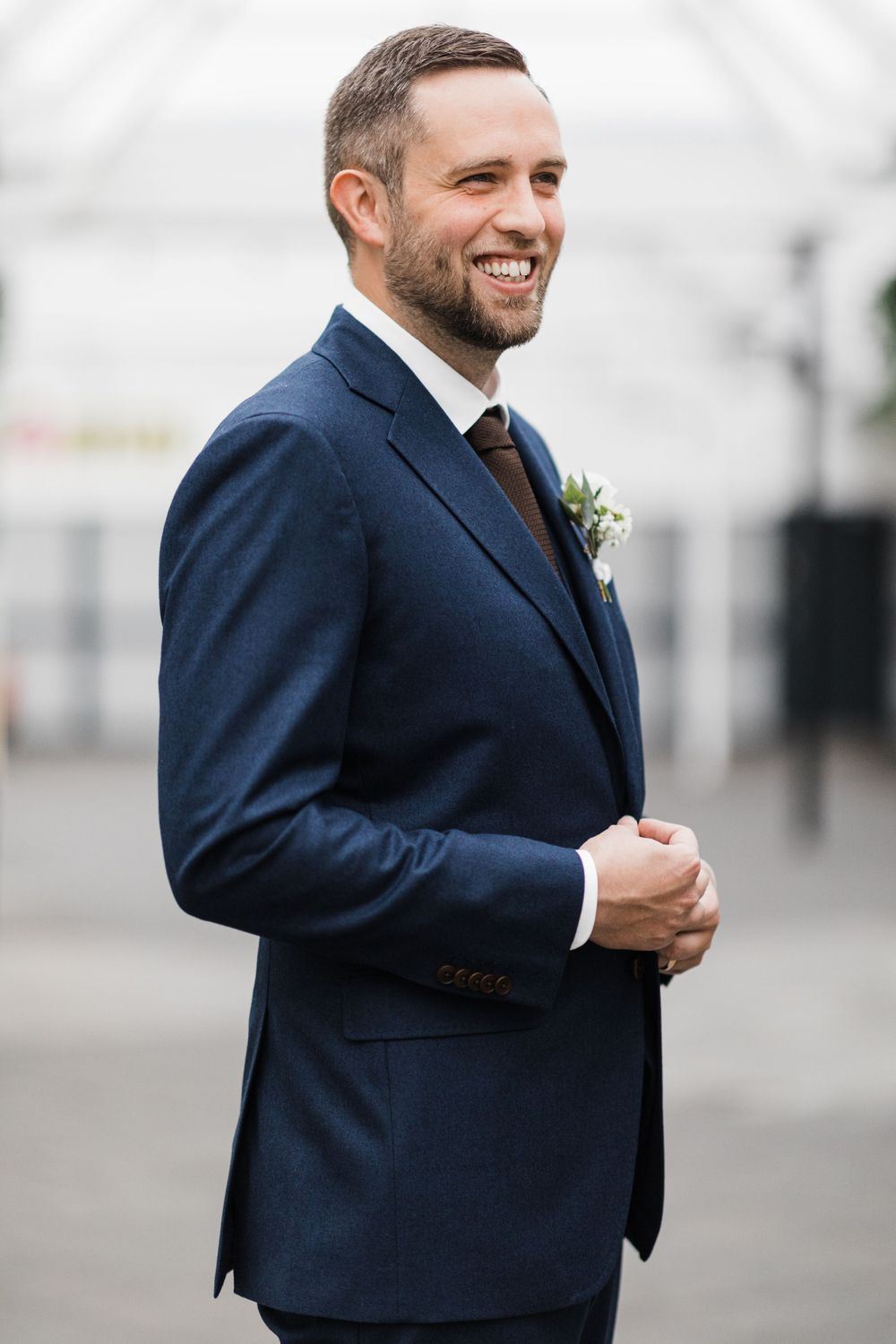 Groom in navy suit with brown tie smiles brightly against light background.