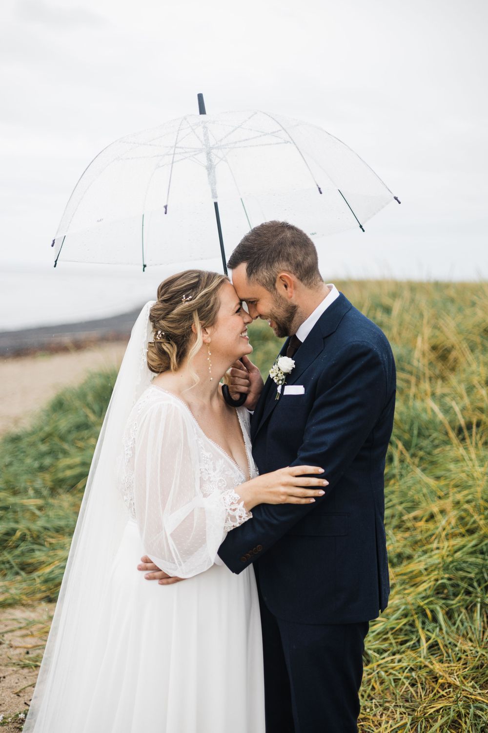 A romantic moment under a clear umbrella against a moody coastal backdrop.